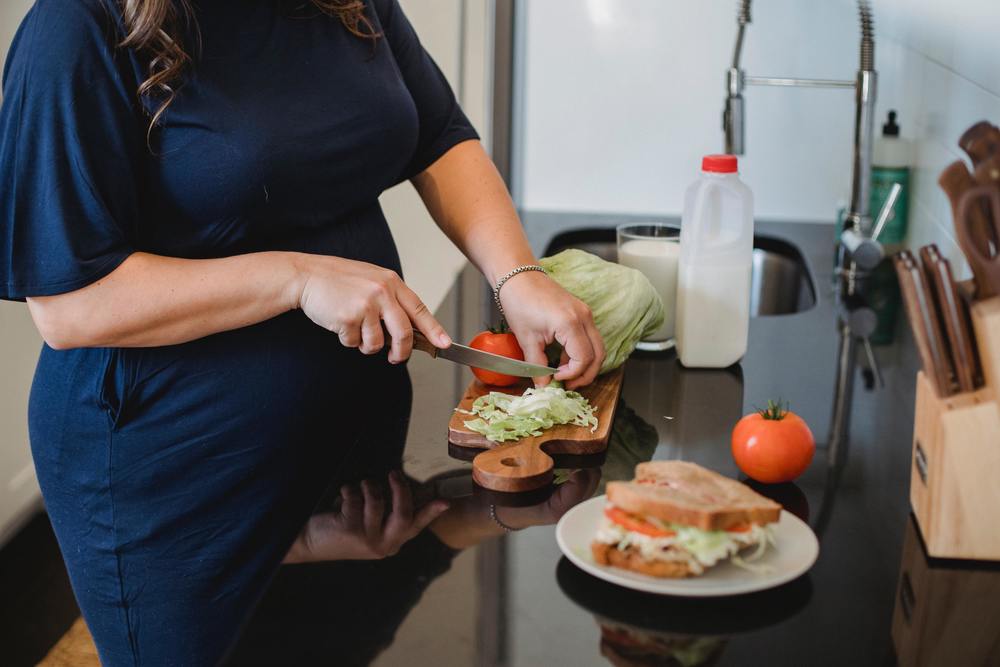 A woman in a navy dress cuts lettuce and tomatoes to assemble a sandwich. A small jug of milk and a full glass sit on the counter in the background.